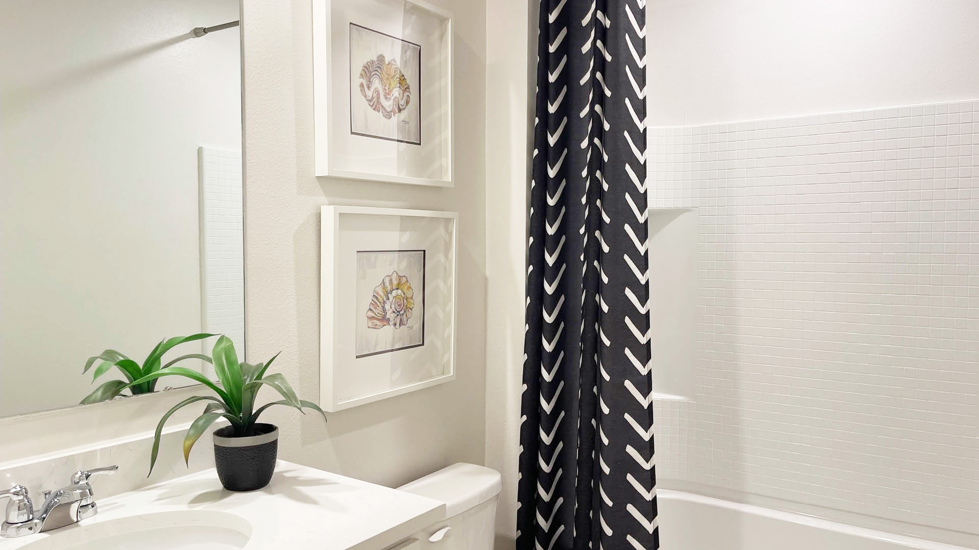 Modern bathroom featuring a white vanity, nautical shell art, a leafy plant, and a patterned shower curtain against light tiles.