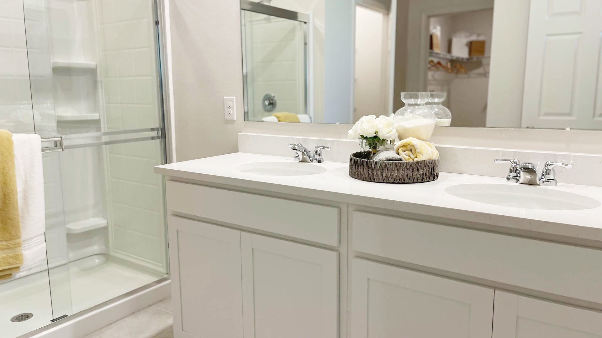 Modern bathroom featuring a double vanity with a quartz countertop, a glass shower, and decorative towels and flowers in a wicker basket.