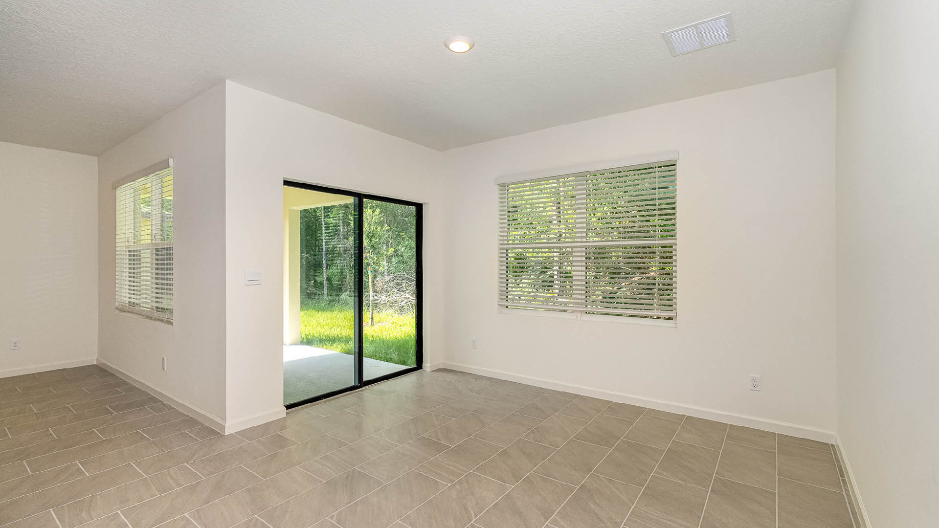 Bright, airy room with large windows and tiled flooring, leading to a green backyard through sliding glass doors.