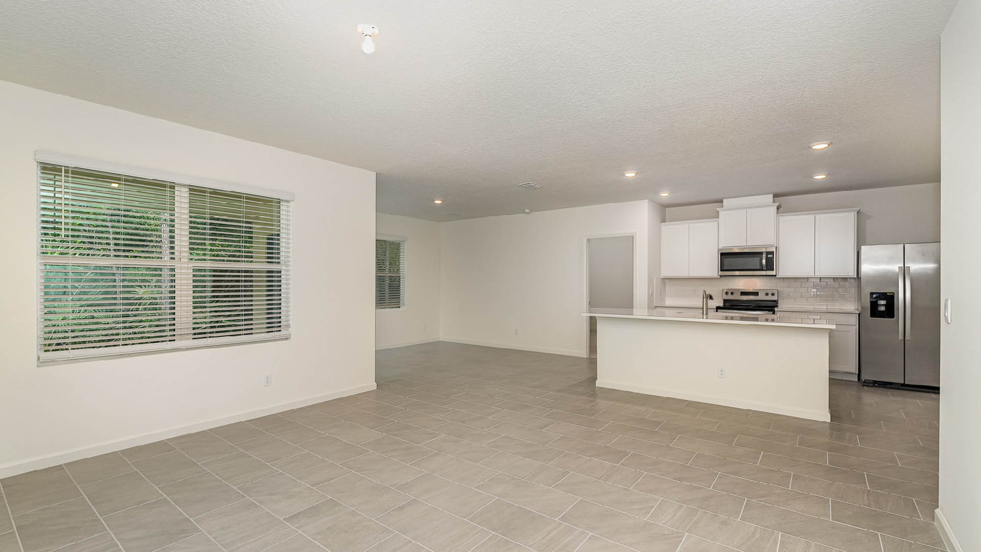 Bright, open-concept living space featuring tile flooring, kitchen with stainless steel appliances, and large windows with blinds.