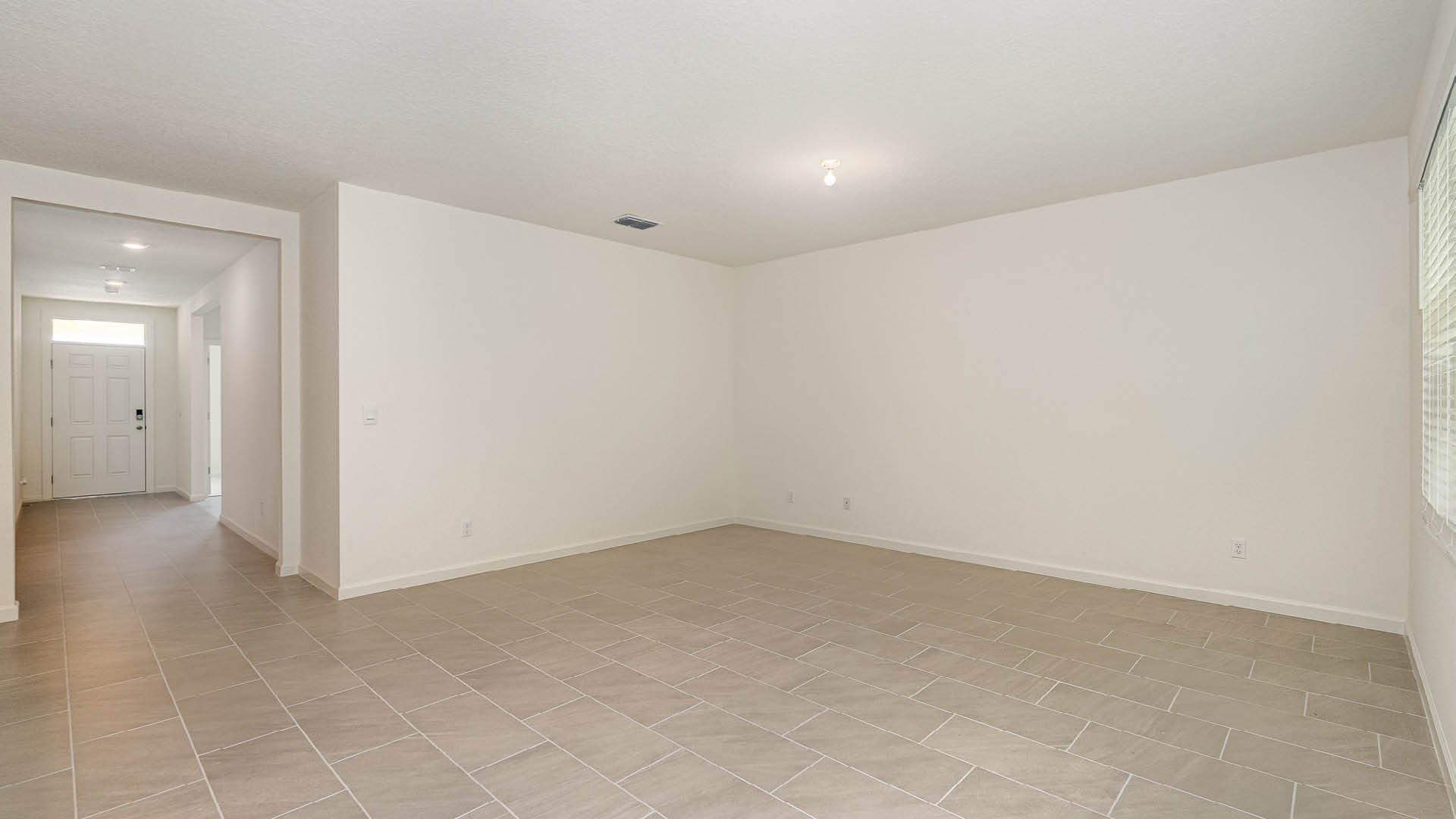 Spacious, empty living area with light tile flooring, neutral walls, and a doorway leading to the entrance. Natural light from windows.