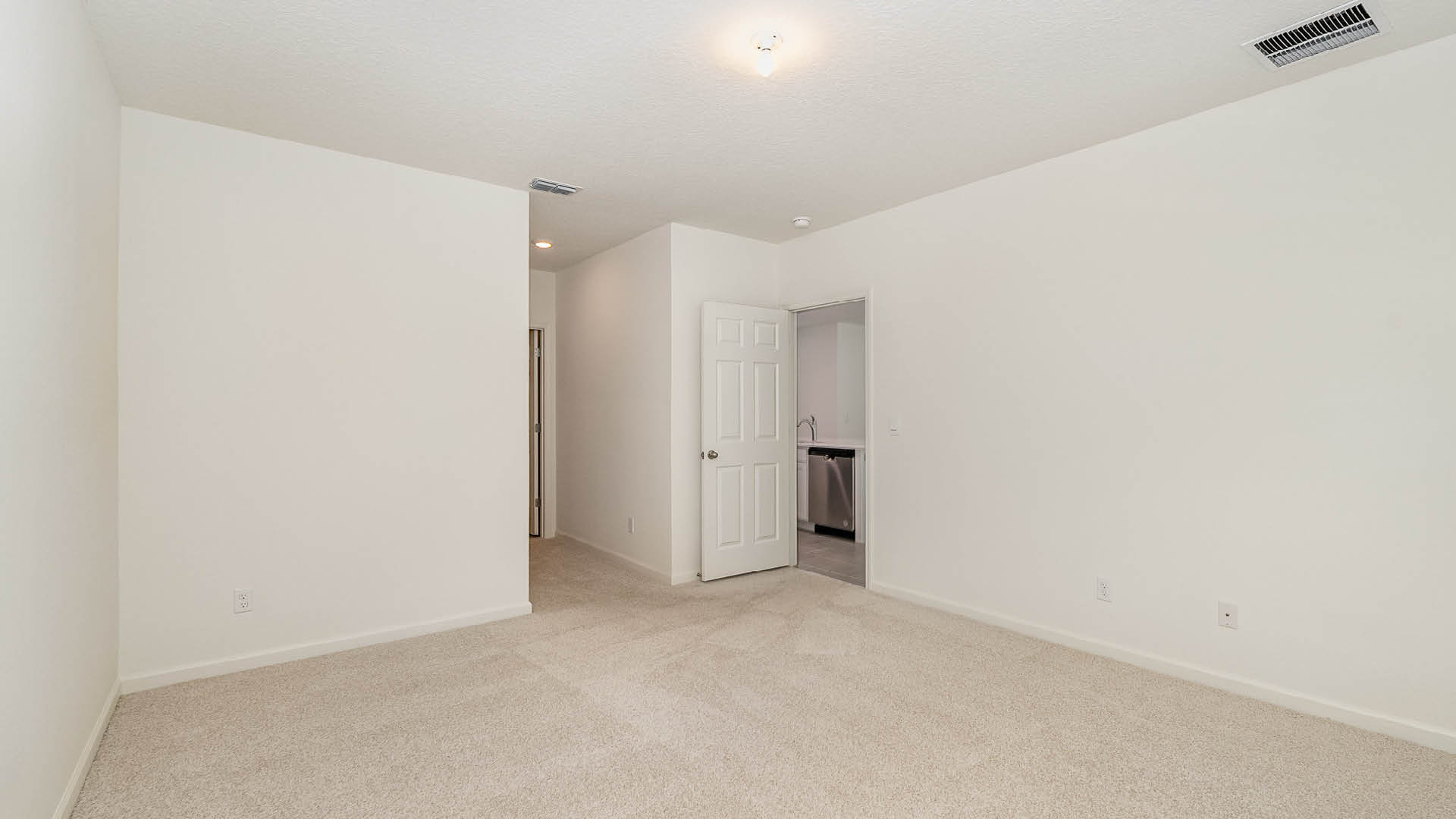 Spacious, empty room with light beige carpet, white walls, and a doorway leading to a kitchen area in the background.