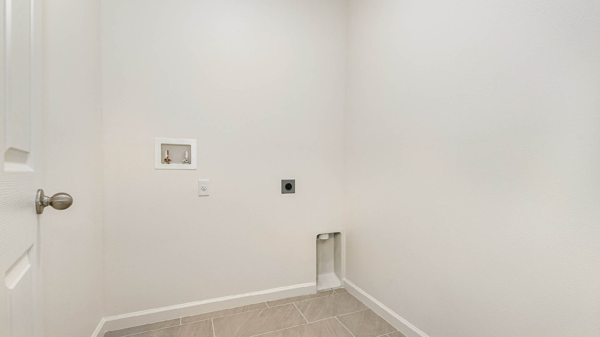 A spacious, empty laundry room with white walls, tiled floor, and utility connections for water and power on one wall.