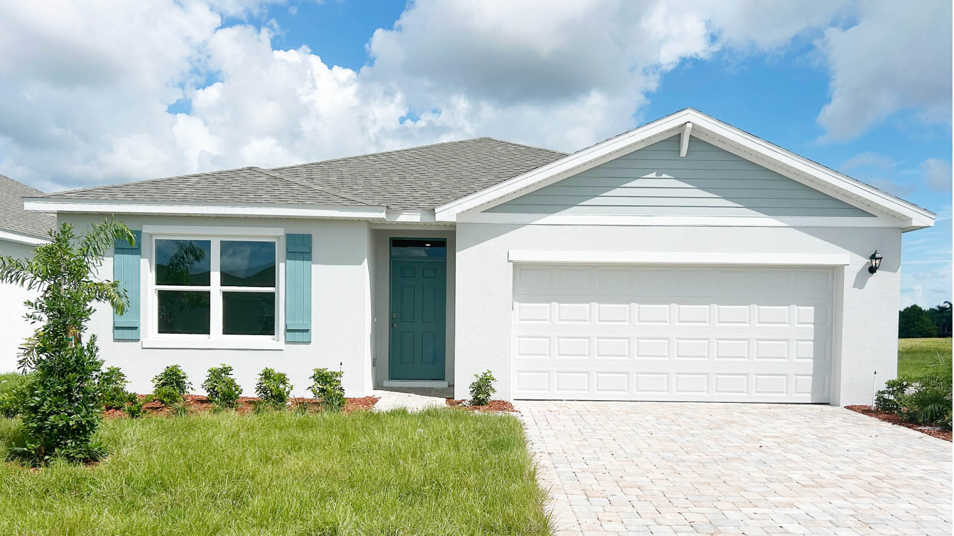 A charming single-story home with a teal front door, light blue shutters, and a paver driveway.