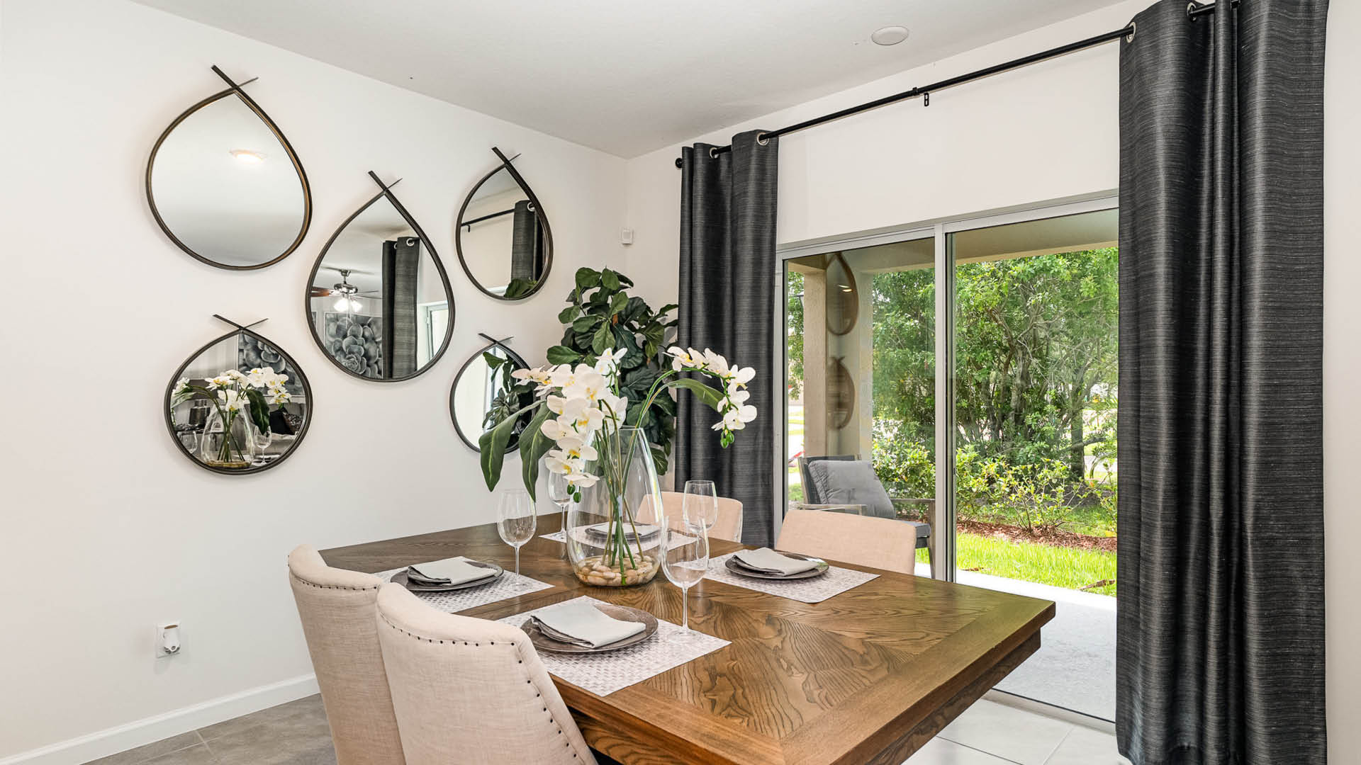 A stylish dining area featuring a wooden table, elegant decor, hanging mirrors, and a view of greenery through sliding glass doors.
