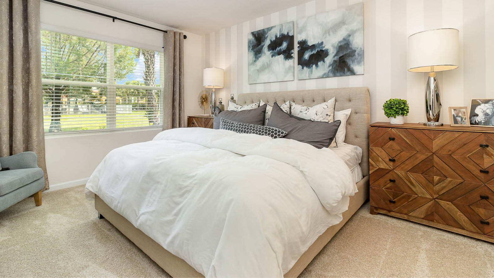 Cozy bedroom featuring a plush bed with layered pillows, a stylish nightstand, and a window with natural light, against soft striped walls.