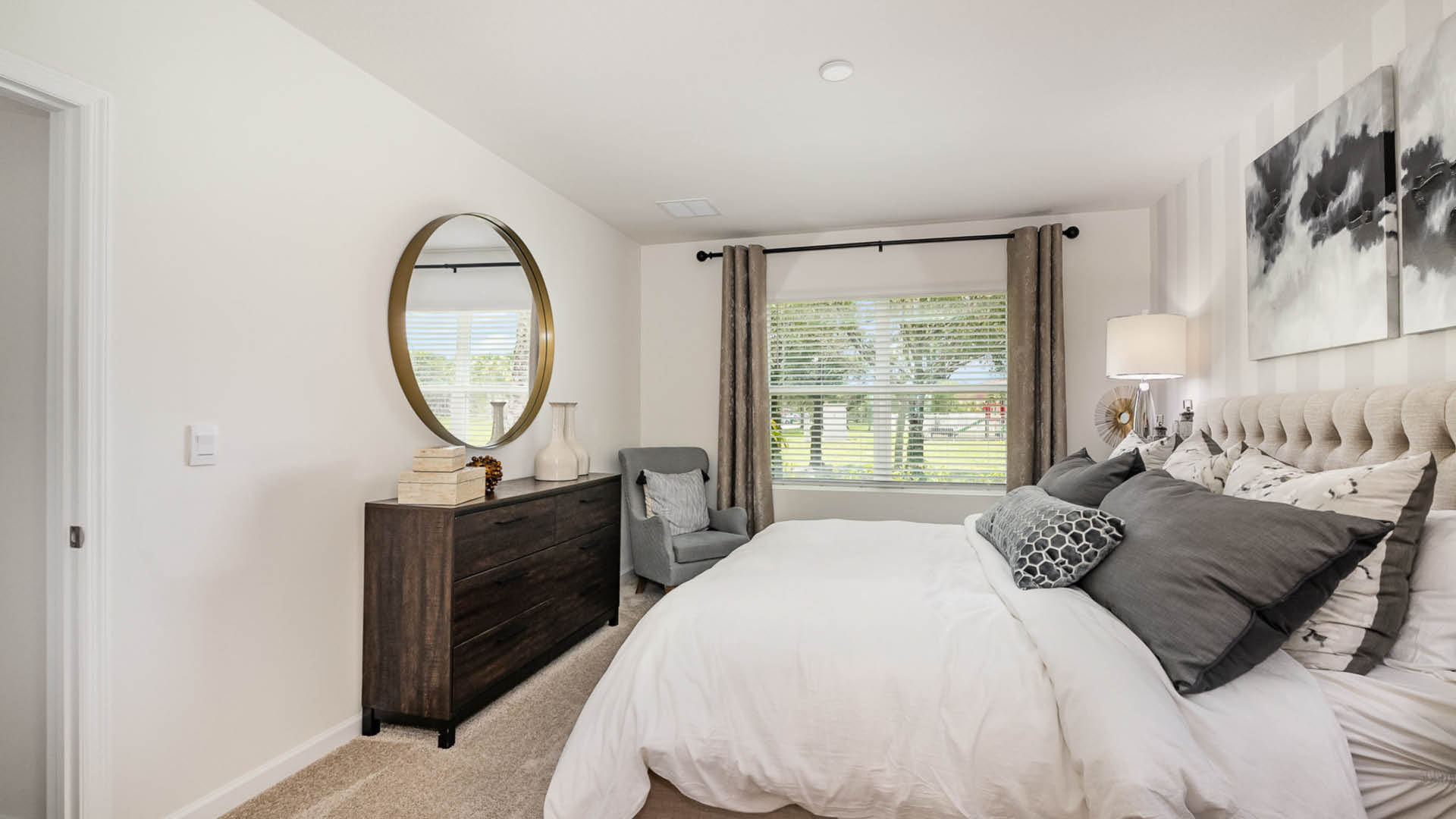 Cozy bedroom featuring a tufted headboard, decorative pillows, a round mirror, and modern furnishings beside a window with natural light.