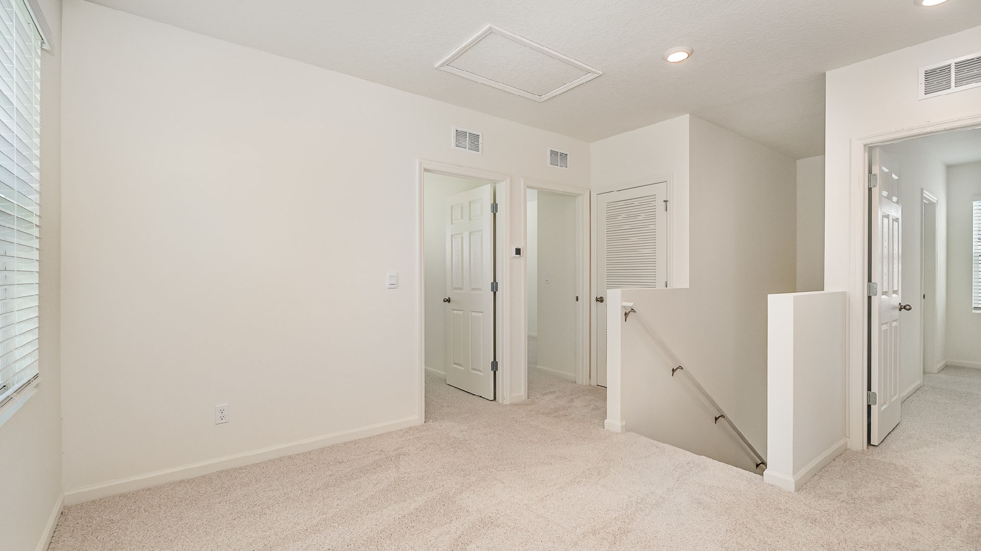 Bright, neutral hallway with plush carpet, featuring doorways to rooms and a staircase, complemented by natural light from a window.