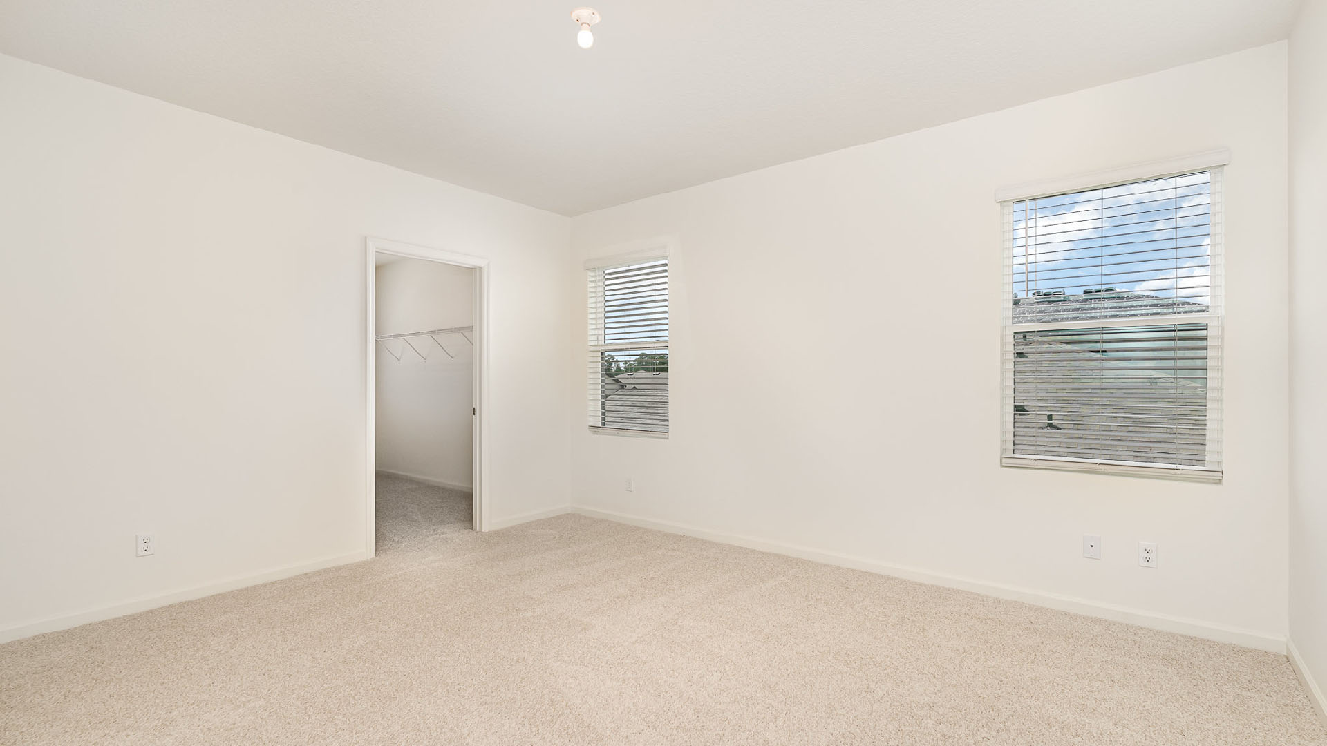 Spacious, empty room with beige carpet, white walls, and two windows with blinds, featuring a closet doorway on the left.