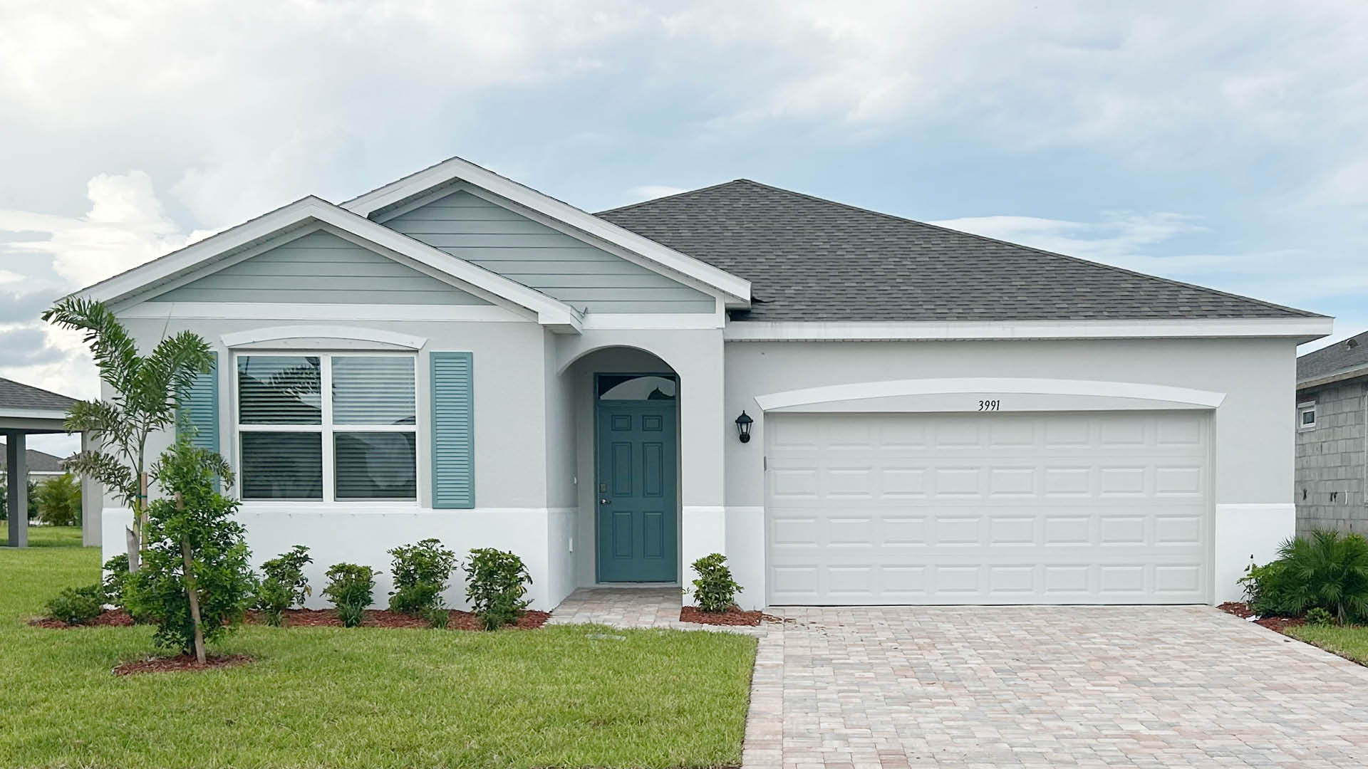 A modern single-family home with a light blue and white facade, a paved driveway, and lush green landscaping in front.