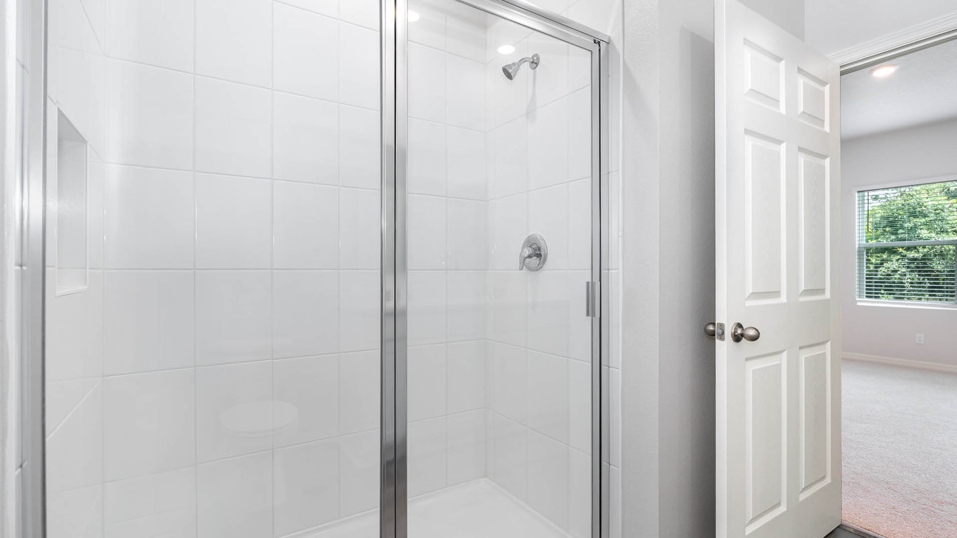 13. Modern glass shower enclosure with white tile and chrome fixtures, adjacent to a doorway leading to a bright, carpeted room.