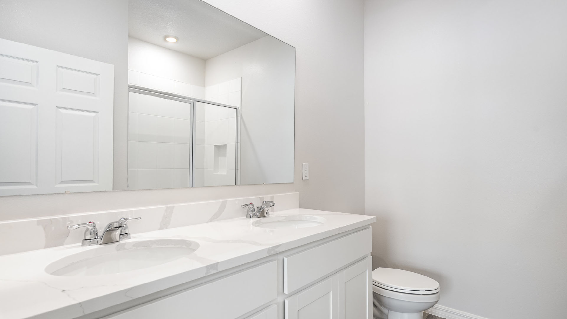Bright modern bathroom featuring a double sink vanity, large mirror, shower, and toilet against a neutral background.