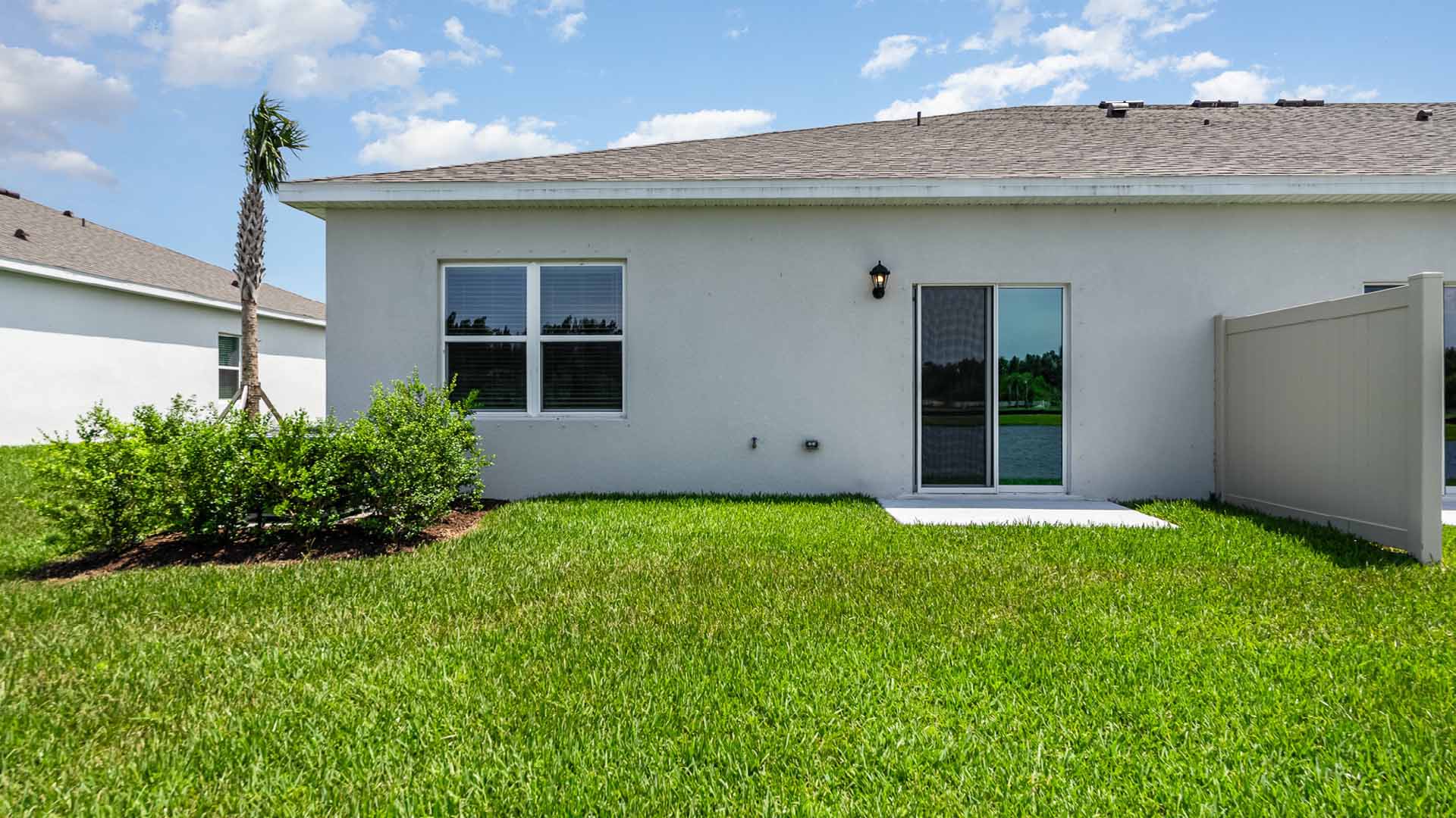A sunny corner of a house featuring a small patio, green lawn, a palm tree, and a gray privacy fence against a blue sky.