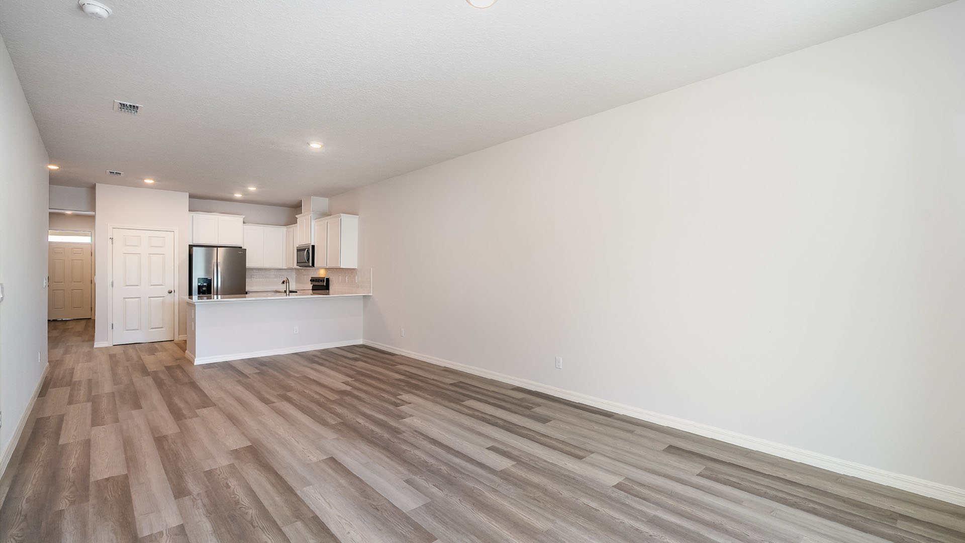 Spacious modern living area with light-colored walls, wood laminate flooring, and a kitchen featuring stainless steel appliances.