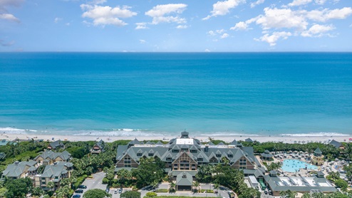 Aerial view of a beachfront resort with a turquoise ocean, cloudy blue sky, and people enjoying the beach and a pool area nearby.