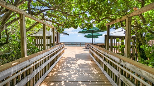 A wooden pathway leads through lush greenery to a sunny deck with umbrellas, overlooking a serene ocean view.