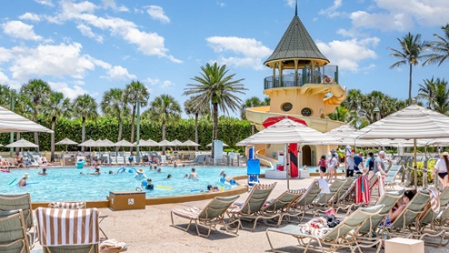 A sunny pool area featuring a water slide, families enjoying the water, and lounge chairs under striped umbrellas amidst palm trees.