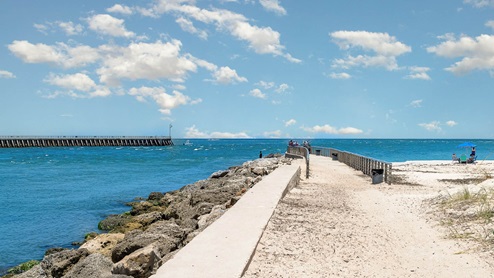 A sandy path leads to a jetty with visitors, all overlooking a vibrant blue ocean under a clear sky with scattered clouds.