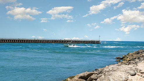 A fishing boat speeds through clear blue waters near a long pier under a sunny sky with clouds. Rocks line the shore.