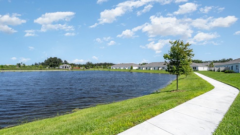 Scenic view of a serene lake surrounded by lush grass and modern homes, with a winding path under a bright blue sky.