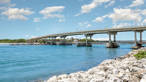 A concrete bridge spans blue waters, with people on the beach below and lush greenery in the background under a clear sky.