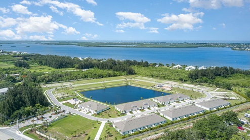 Aerial view of a residential community with homes surrounding a central pond, near a large water body and lush greenery.