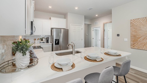 Modern kitchen featuring white cabinets, stainless steel appliances, and a sleek island with quartz countertops and dining setup. Neutral decor and bright lighting.