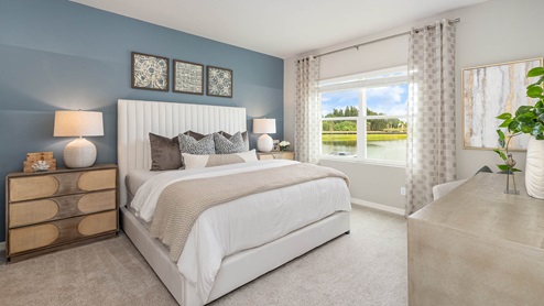 A bedroom with a large bed, decorative pillows, bedside lamps, and artwork on the wall. A window shows a view of a lake and trees. A dresser with plants is on the right.