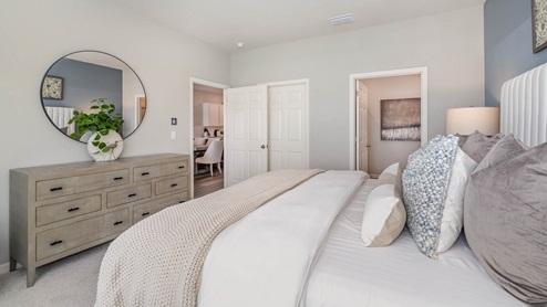 Bright bedroom featuring a large bed with decorative pillows, modern dresser, round mirror, and an open doorway to a kitchen area.