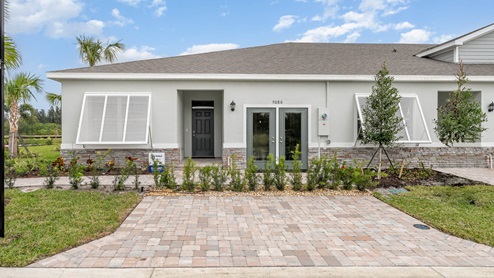 A modern, gray villa with stone accents, two front doors, paver driveway and landscaped garden, set against a blue sky and green surroundings.
