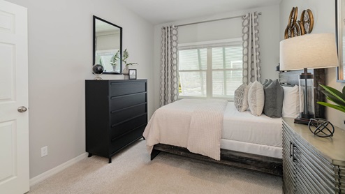 Cozy bedroom featuring a bed with decorative pillows, a black dresser, a stylish lamp, and a mirror on the light gray walls