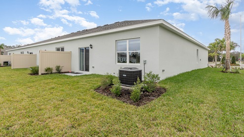 Side view of a modern white home with a grassy yard, an air conditioning unit, and a privacy fence, under a blue sky.