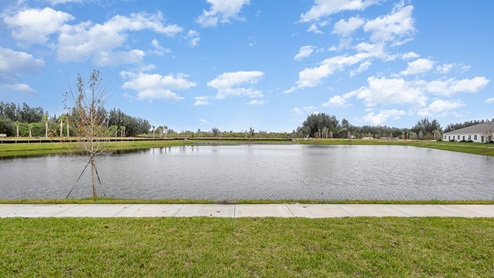 A serene view of a lake surrounded by lush greenery and a paved walkway under a clear blue sky with scattered clouds.