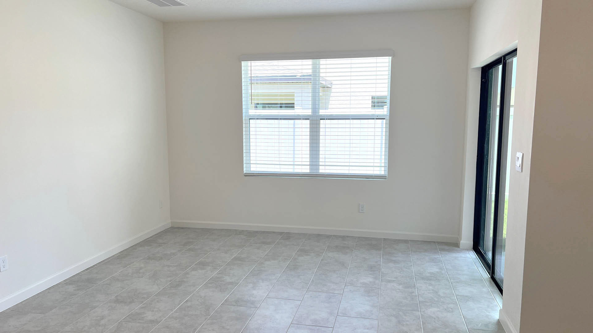 Bright, empty room with light gray tile flooring, a large window with blinds, and sliding glass doors leading outside.