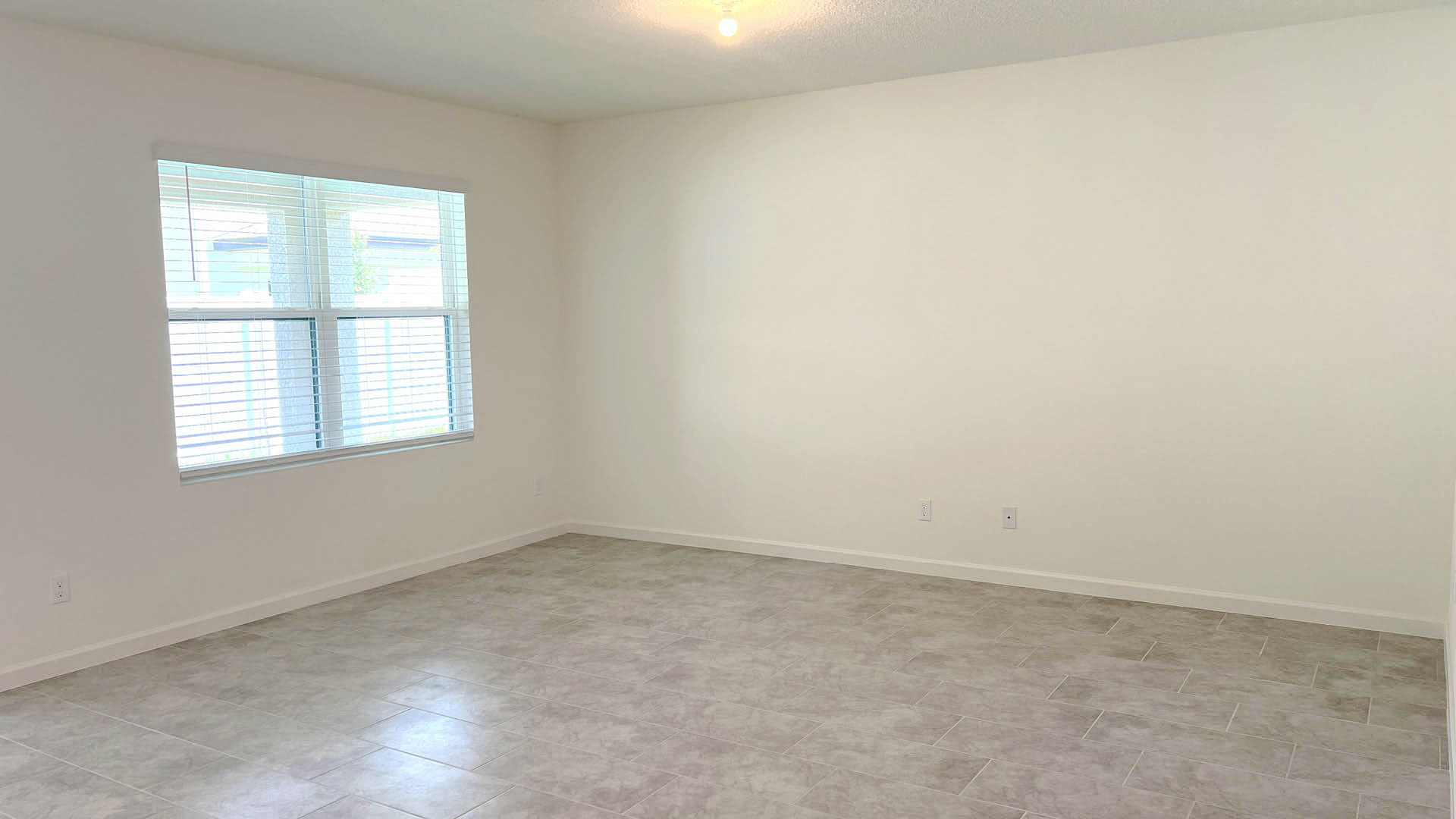 Spacious, empty room with gray tiled flooring, a large window with blinds, and white walls illuminated by overhead lighting.