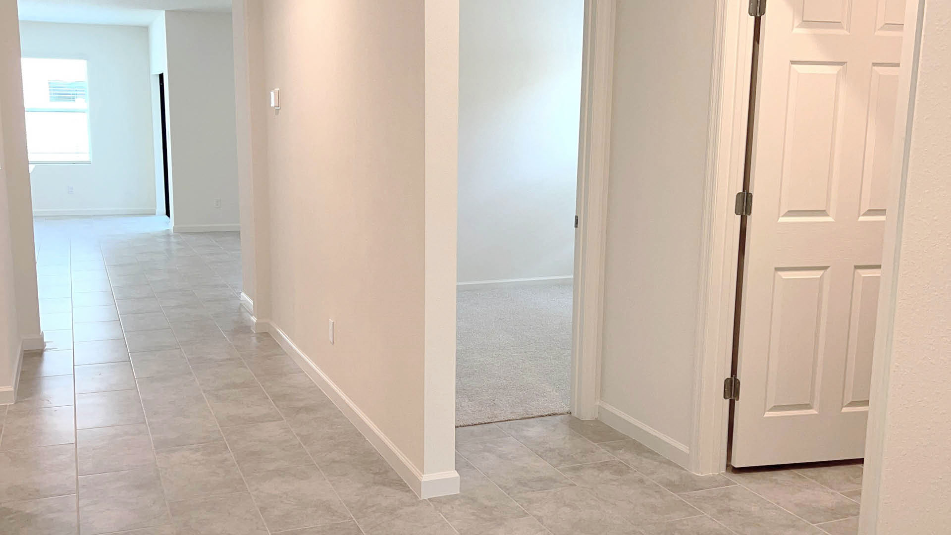 Bright hallway with tiled floor leading to doorways, one opening into a carpeted room. Light walls enhance the spacious feel.
