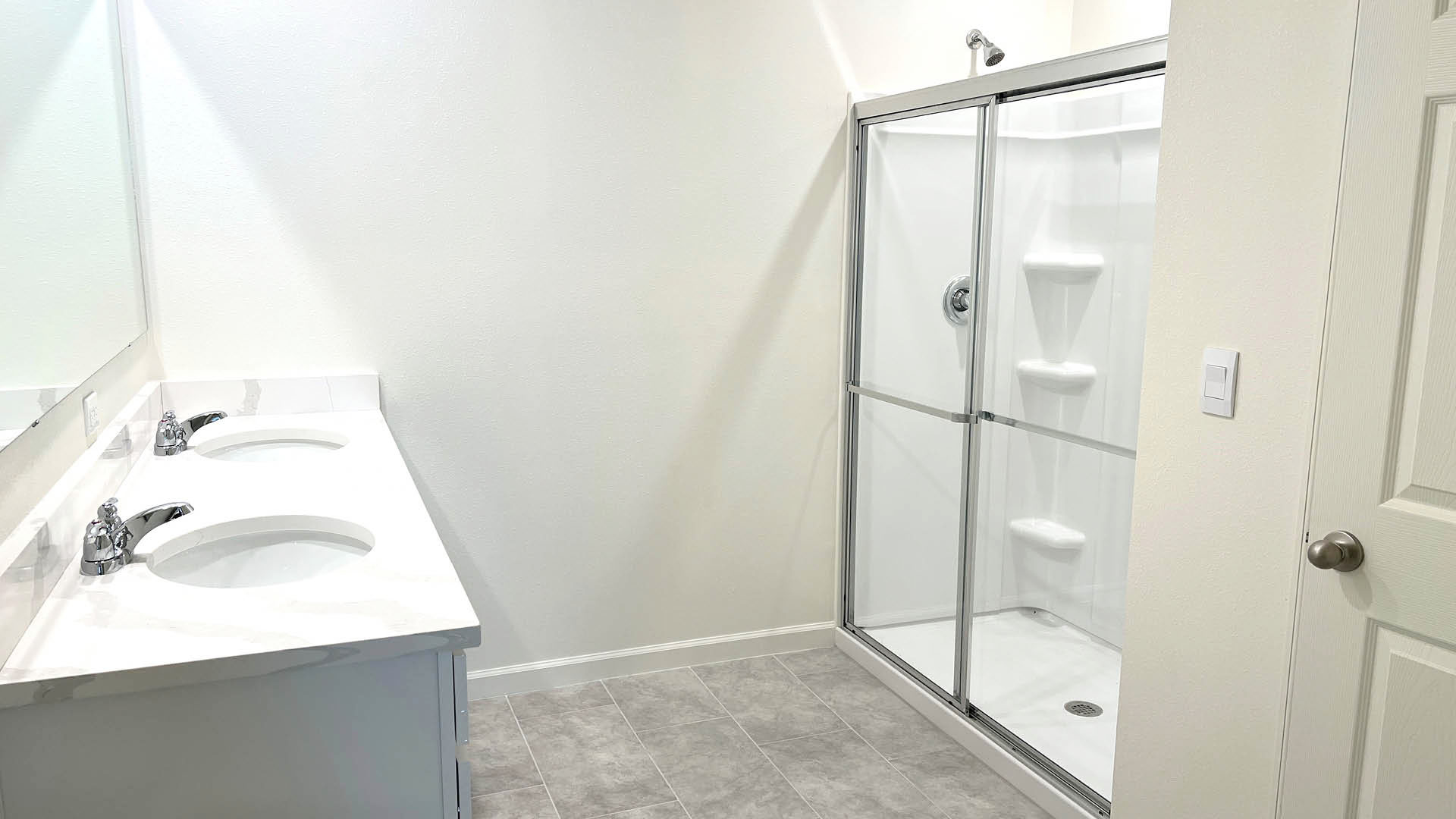 Bright bathroom featuring dual sinks with a quartz countertop, a glass-enclosed shower, and light gray tiled flooring.