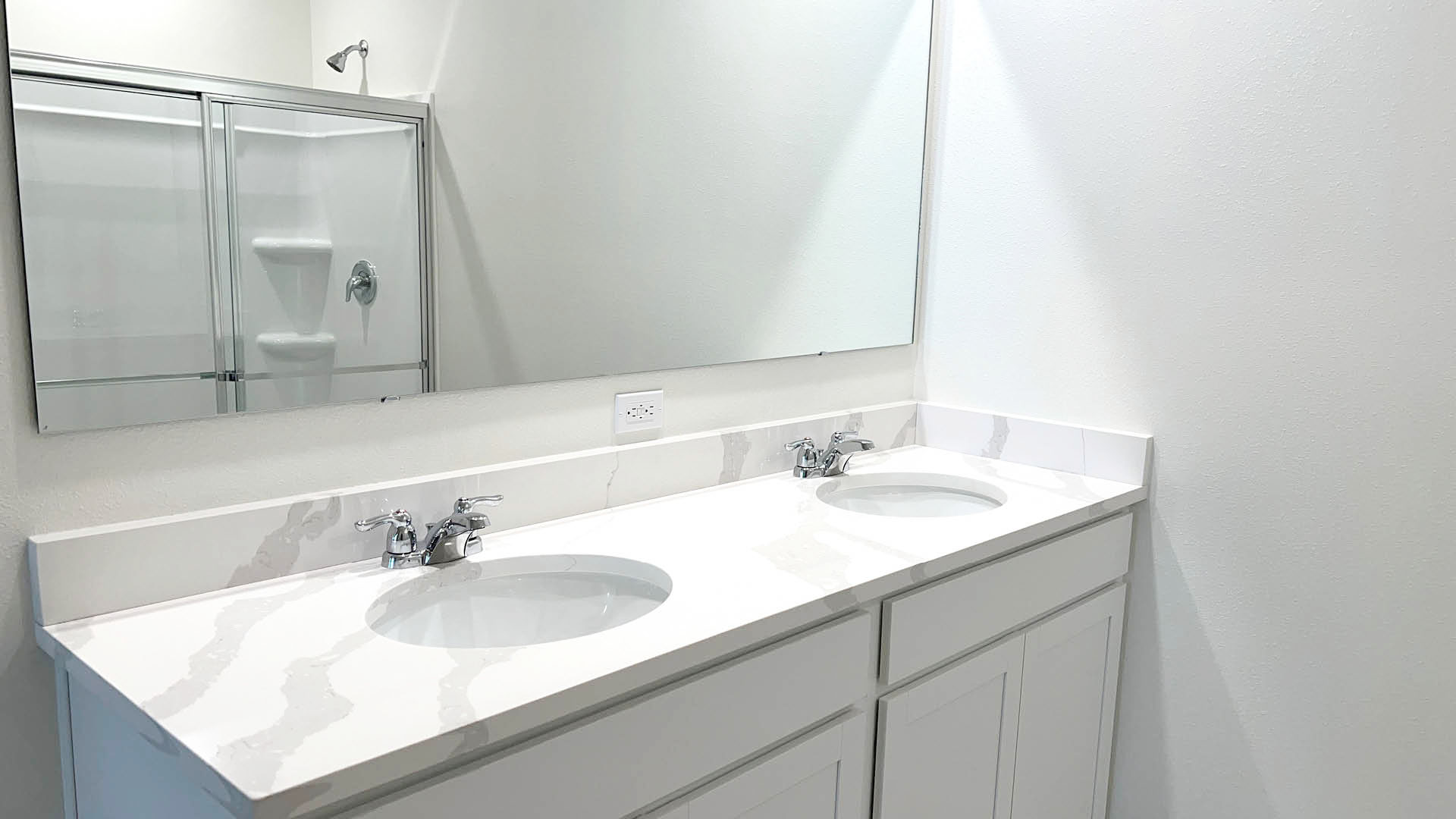 Modern bathroom with double sinks, white quartz countertop, and a large mirror, featuring a glass shower enclosure in the background.
