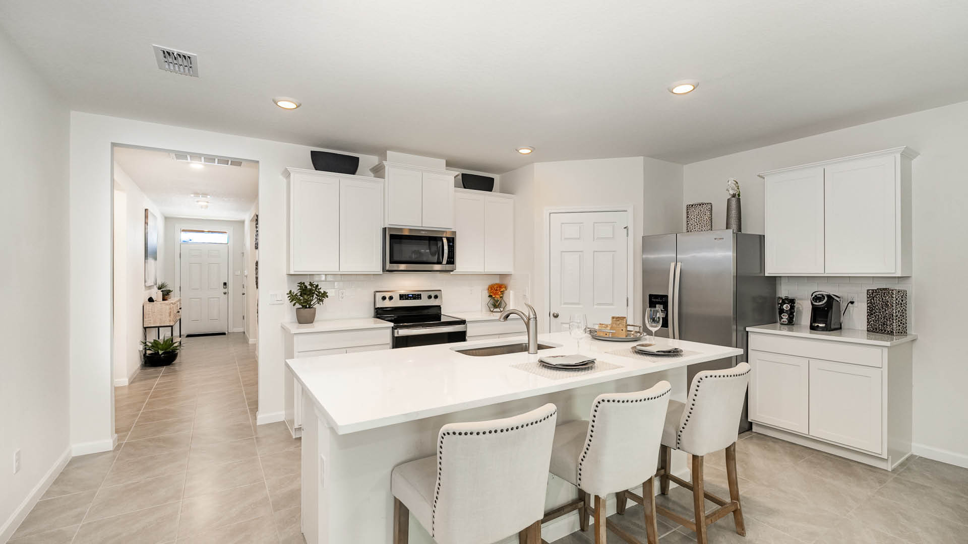 Bright, modern kitchen with white cabinetry, stainless steel appliances, and a spacious island with bar stools, leading to an entryway.