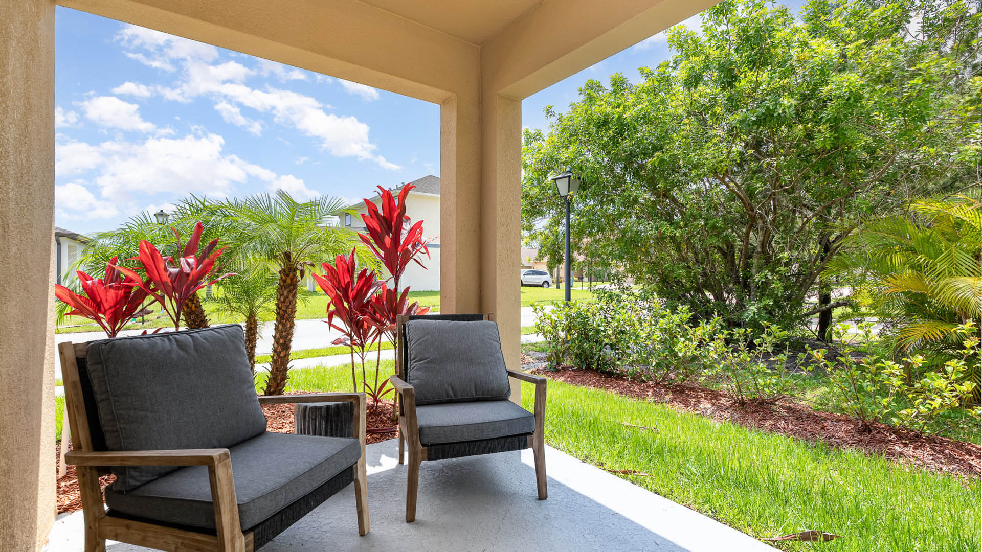 A cozy porch scene featuring two gray cushioned chairs, vibrant red foliage, and lush greenery.