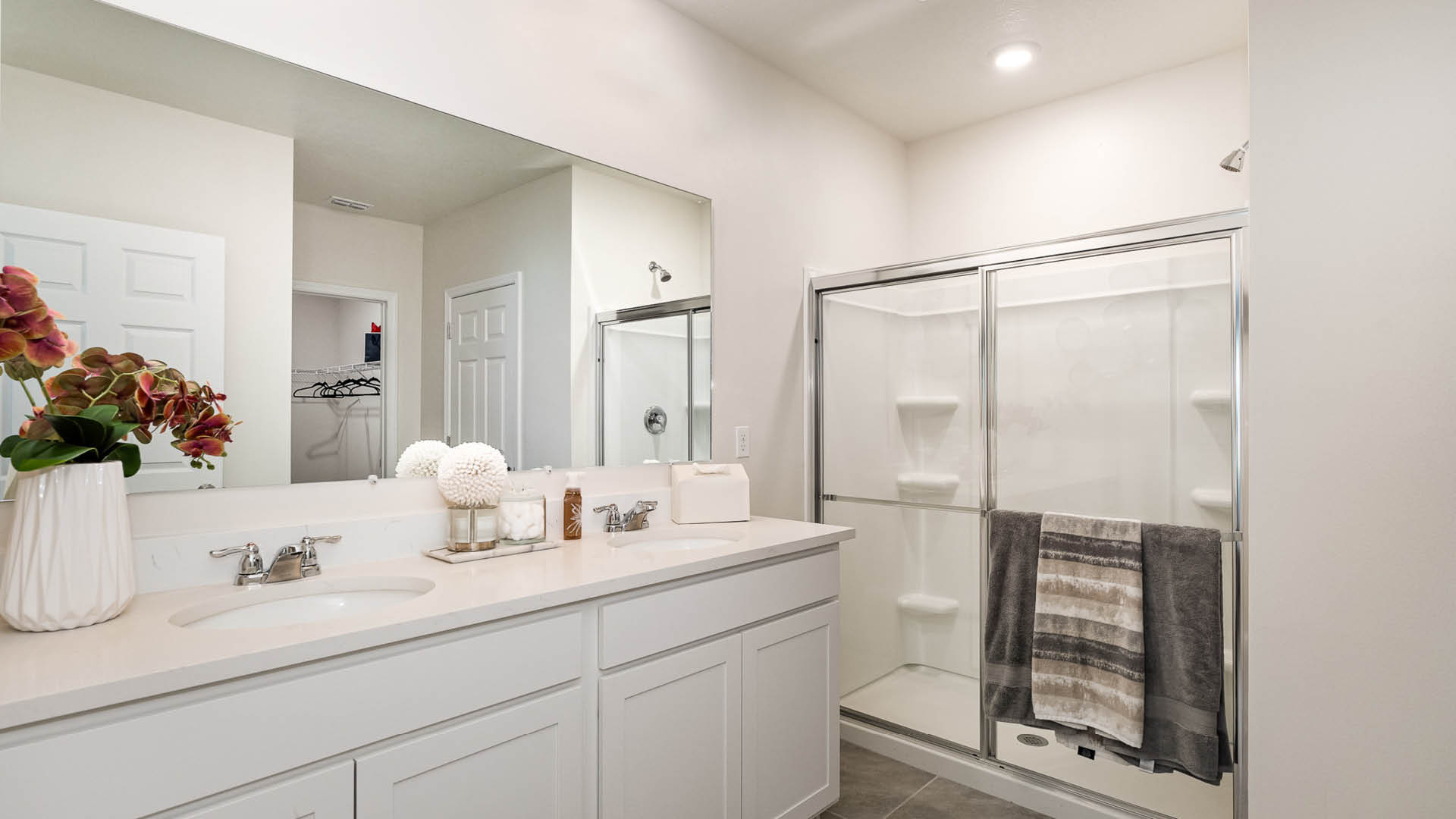 Modern bathroom featuring a double sink vanity, glass shower, neutral decor, and a mirror reflecting a spacious closet.
