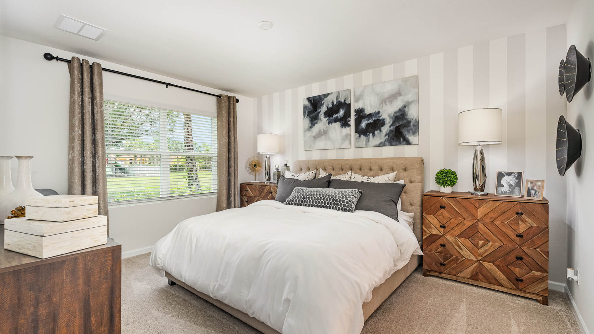 Cozy bedroom featuring a tufted bed with gray and white bedding, decorative pillows, wood accents, and window with landscaped view.