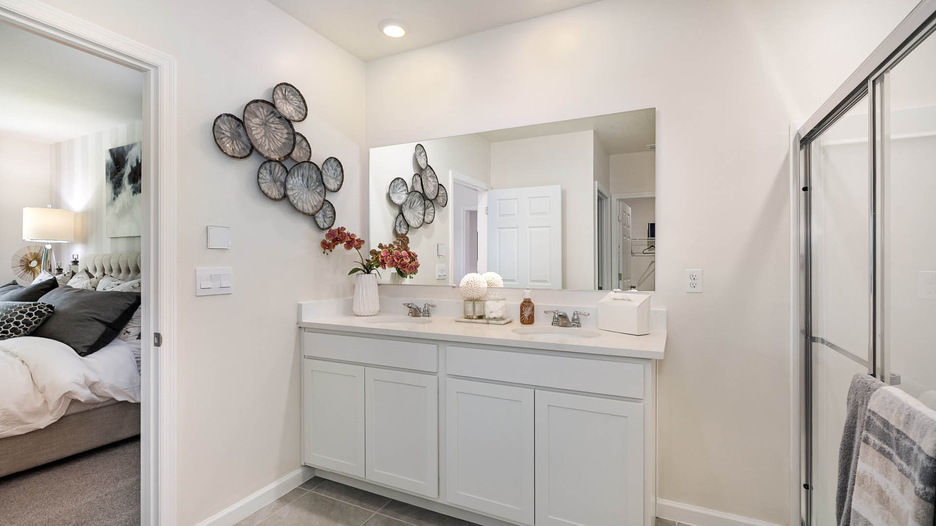 Bright, modern bathroom featuring a double vanity, decorative wall art, and a glass shower, connected to a cozy bedroom.