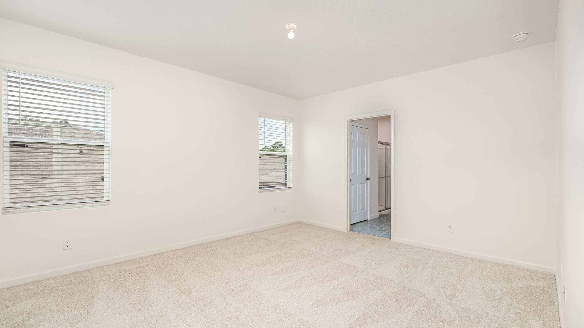 Bright, empty bedroom with beige carpet and white walls, featuring two windows with blinds and an open door leading to a bathroom.