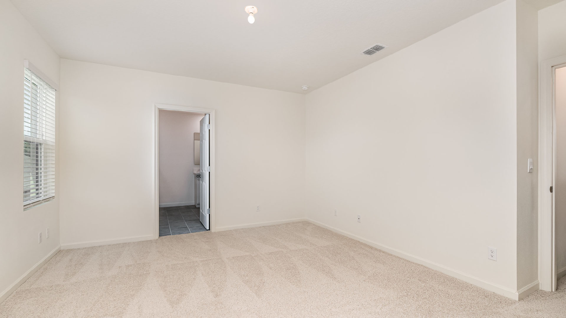 Spacious, empty room with beige carpet, white walls, a window, and an open door leading to a laundry area. Soft natural light filters in.