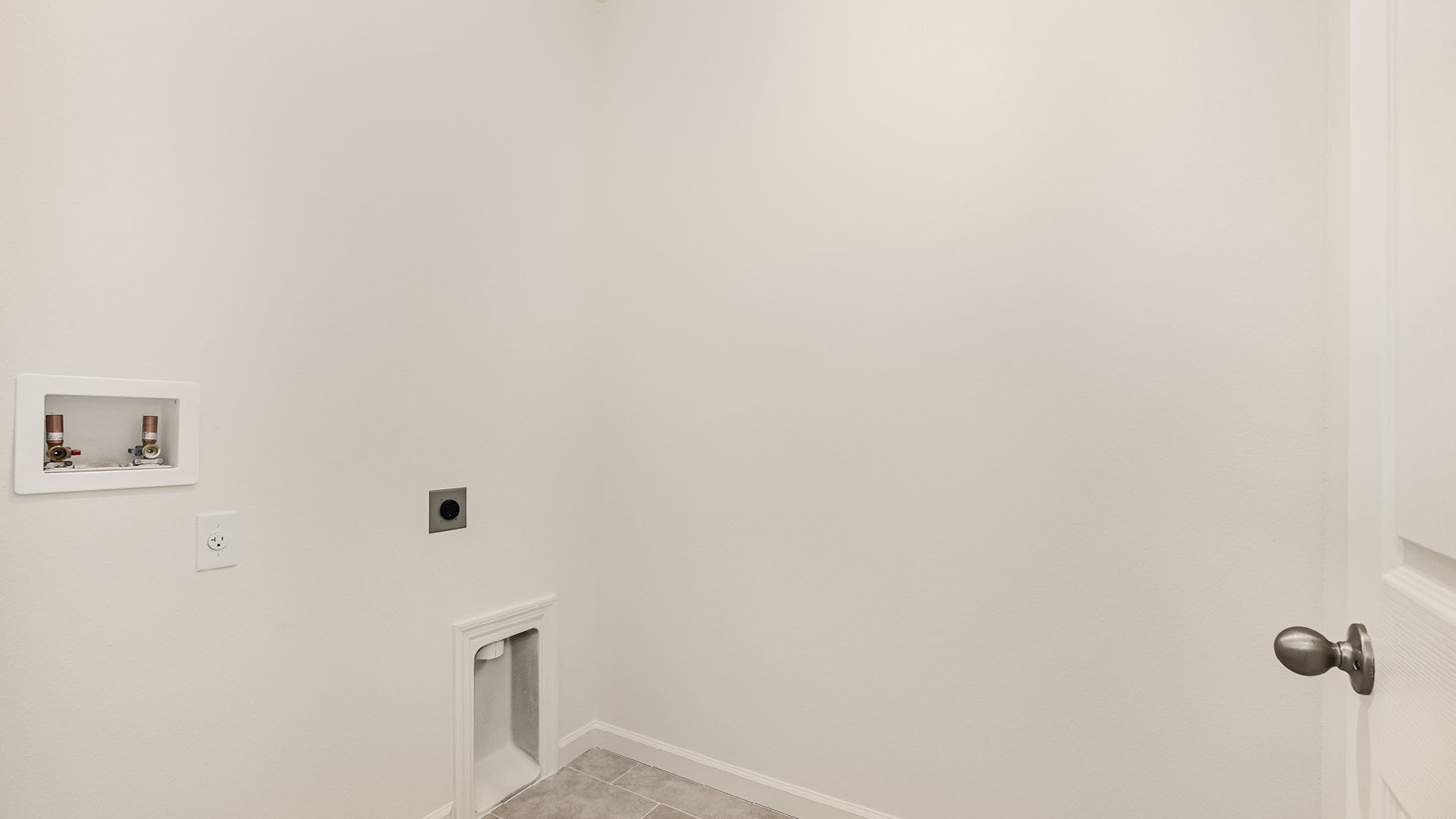 Interior of a laundry room featuring white walls, tiled floor, and utility connections, with an empty corner and a door handle visible.