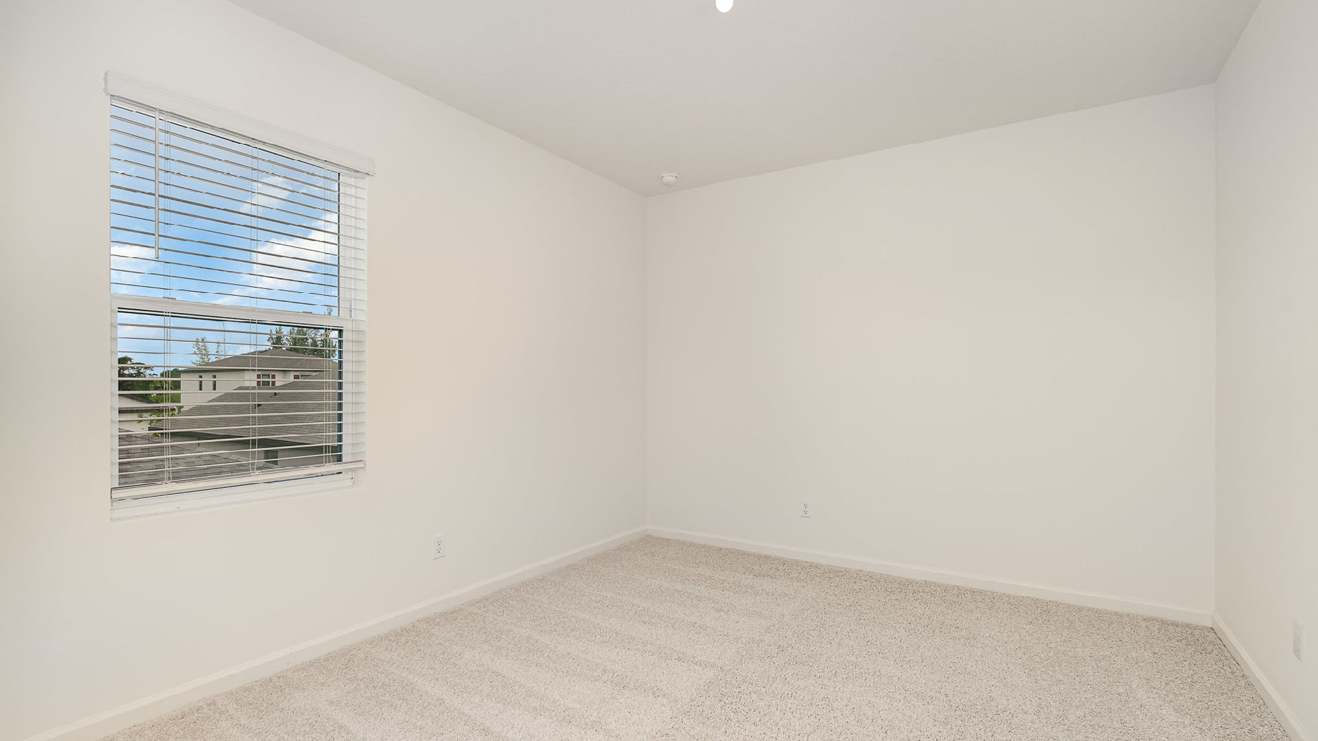 Bright, empty room with light-colored carpet, a window with blinds, and soft natural light illuminating the white walls.