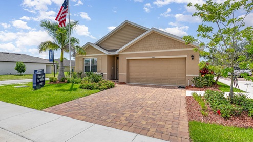 A charming single-story home with a tan exterior, lush landscaping, a flagpole, and a welcoming driveway