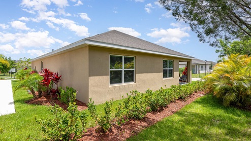 Side view of a single-story home with lush greenery, colorful plants, and a clear blue sky, creating a welcoming outdoor space.