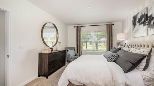 Cozy bedroom featuring a tufted bed, stylish nightstand, a large round mirror, and a window with sheer curtains overlooking greenery.
